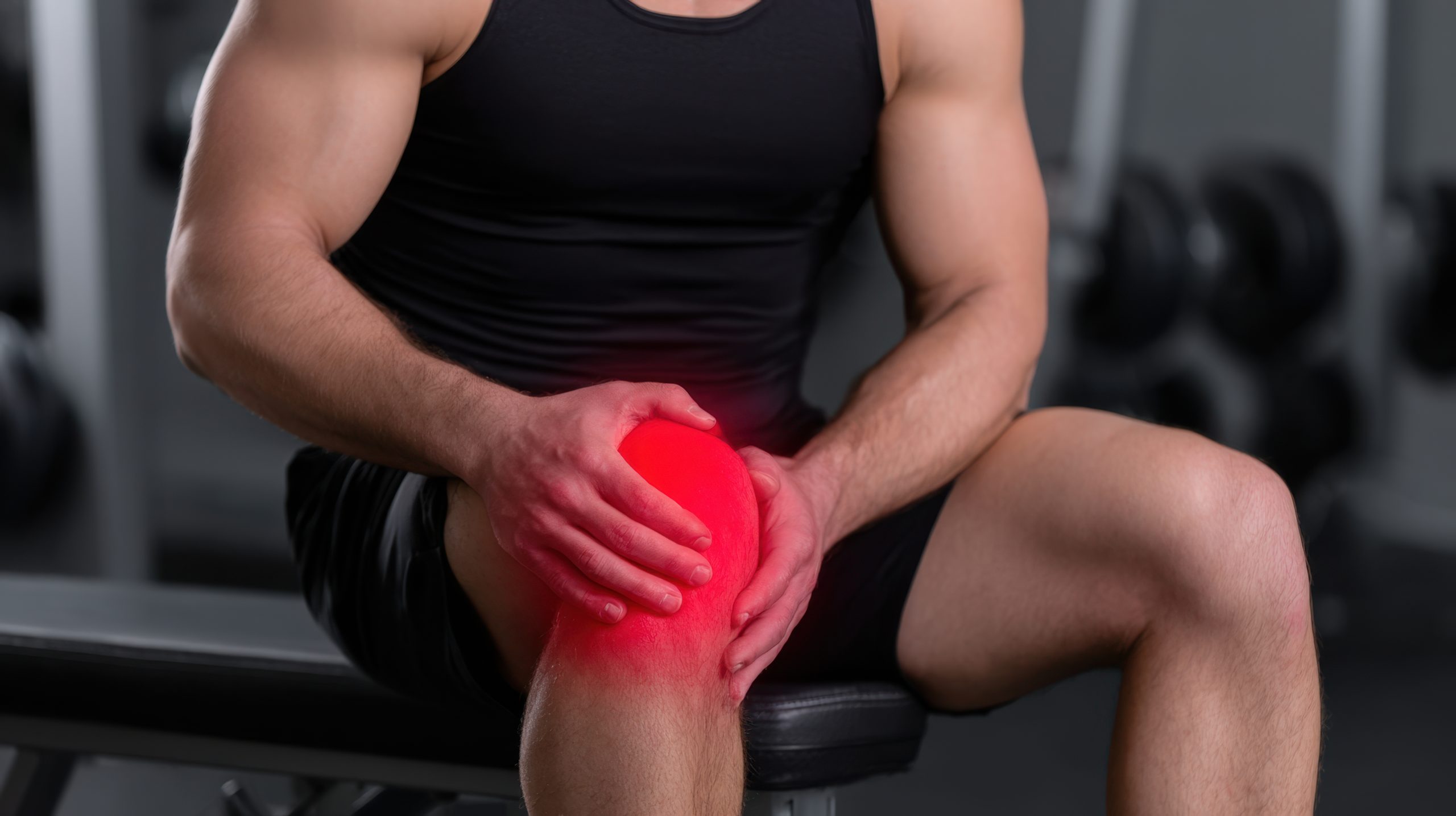 Person experiencing knee pain during workout session in a gym environment with weights in background